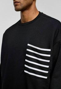 Zwarte sweatshirt met een gestreepte stofzak met horizontale witte strepen. Zachte textuur, ronde halslijn en geribde manchetten.