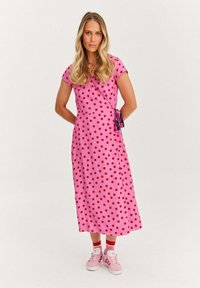 Růžové šaty s puntíky v červené barvě, zavinovací design, krátké rukávy, uvázané na boku. Nošeny s červenými ponožkami a růžovými teniskami. Hladká textura.