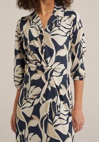 Blumenkleid mit einer Farbkombination aus Marineblau und Creme, mit kragenförmigem Ausschnitt, Dreiviertelärmeln und einem gedrehten Vorderdesign. Texturierter Stoff.