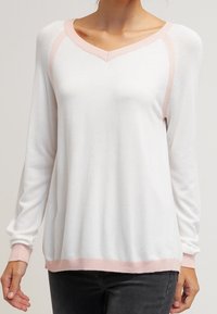 Donna che indossa un maglione bianco a maniche lunghe con rifiniture rosa chiaro su colletto, polsini e orlo, abbinato a pantaloni scuri.