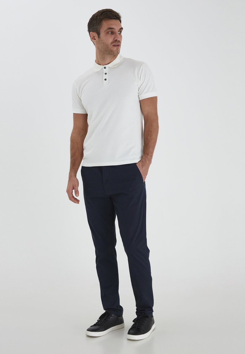 White polo shirt with black buttons and short sleeves, paired with navy blue trousers and black sneakers. Simple design and fitted silhouette.