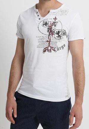 Man draagt een wit henley-shirt met korte mouwen met zwarte en rode heraldieke ontwerpen en tekst, gecombineerd met donkere broek.