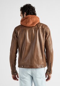 Bruine leren jas met een slim fit ontwerp, voorzien van een oranje capuchon, ritsmanchetten en zichtbare stikseldetails. Lichte spijkerbroek zichtbaar eronder.