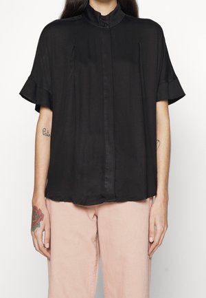 Femme portant un chemisier noir ample à manches courtes avec un col montant et un pantalon rose pâle, montrant des tatouages sur ses bras et sa main.