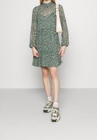 Vestido floral verde con mangas largas y transparentes, un dobladillo con volantes y un escote alto. Combinado con zapatillas blancas y calcetas de rayas rosas.