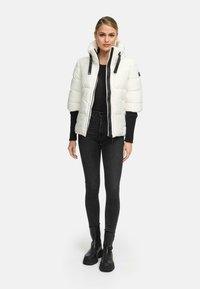 Vrouw in een witte pufferjack met korte mouwen over een zwart lang shirt, zwarte skinny jeans en zwarte enkellaarsjes.