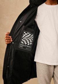 Čierna bunda s jemnou fleecovou podšívkou, ktorá má na vnútornej strane vrecka kostkovanú nášivku loga. Vonkajší materiál je hladký.