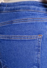 Tela de mezclilla azul con un bolsillo trasero, con costuras visibles en un color marrón claro contrastante. Textura suave y diseño clásico de jeans.
