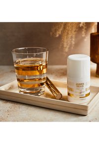 Un bicchiere di vetro trasparente con strisce dorate contiene un liquido, accanto a una bottiglia bianca con tappo dorato etichettata "JAMES READ Gradual Tan" su un vassoio beige.