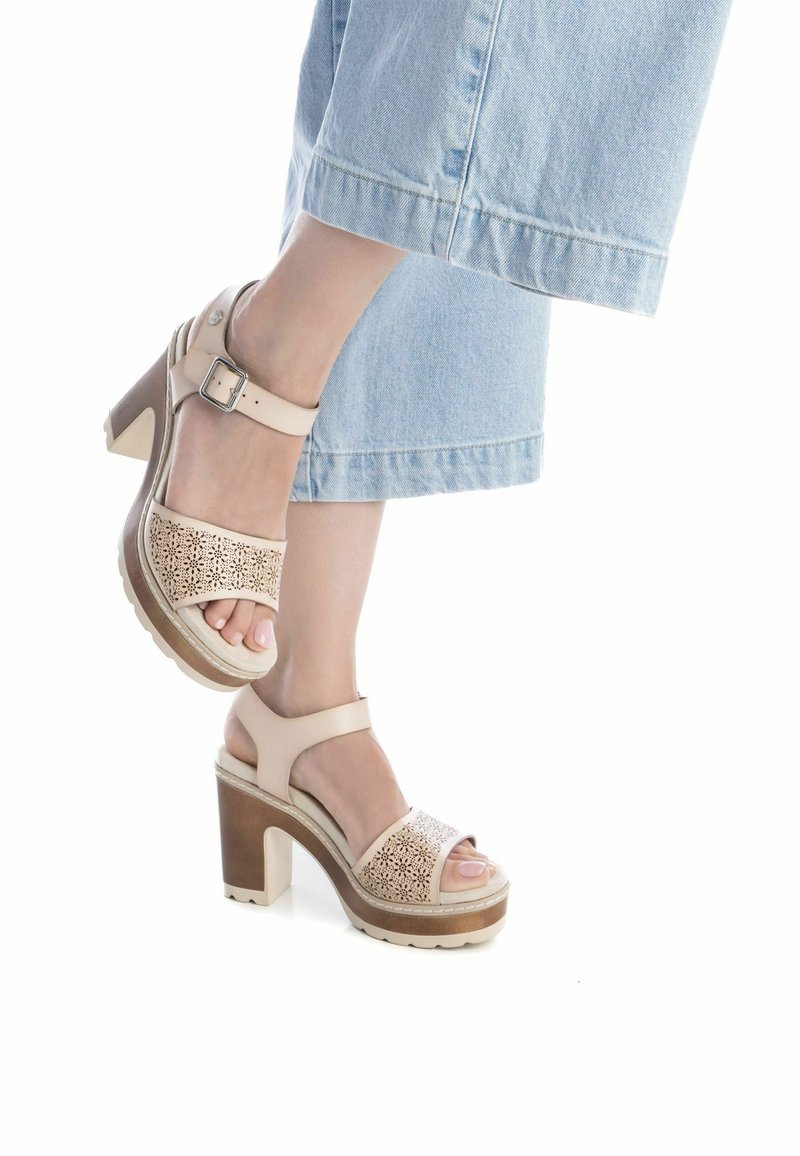 Sandalias de tacón alto en color beige con un diseño recortado, tira en el tobillo y tacón de madera. La suela es gruesa y texturizada para ofrecer agarre.