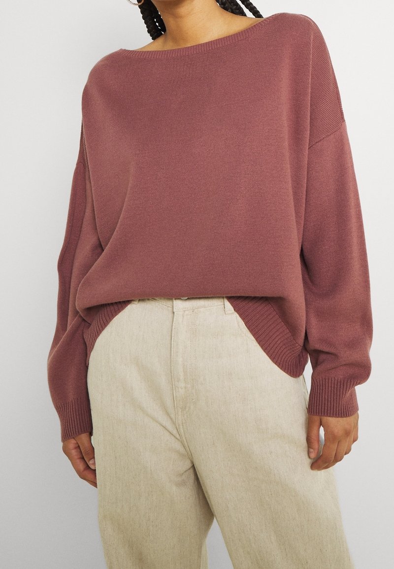 Femme portant un pull ample mauve à manches longues et un pantalon couleur crème, montrée des épaules aux hanches sur un fond uni.