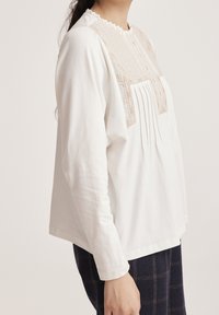 Top blanco de manga larga con diseño plisado y bordado detallado en el escote, con un corte holgado y dobladillo redondeado.