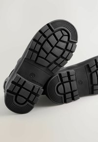 Semelles de chaussures en caoutchouc noir avec un motif de semelle profond, comportant des rainures circulaires et rectangulaires pour une meilleure adhérence. Indication "Fabriqué en Chine" et taille 12 marquées.
