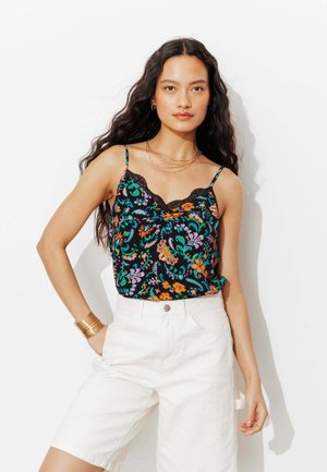 Femme aux longs cheveux foncés portant un caraco noir orné de dentelle florale et un short blanc taille haute avec des bijoux dorés sur un fond blanc.