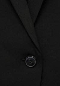 Black blazer fabric close-up, featuring a textured surface and a round, black button detail on the lapel.