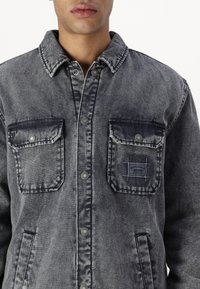 Chaqueta de mezclilla en gris desgastado con cierre de botones, dos bolsillos en el pecho y una etiqueta en el bolsillo izquierdo. Cuenta con un cuello clásico y una textura resistente.
