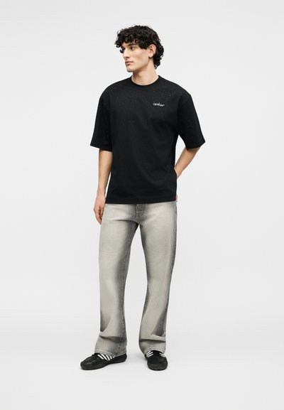 Νέος άνδρας με σγουρά μαλλιά, φορώντας μαύρο κοντομάνικο πουκάμισο, ξεθωριασμένο γκρι τζιν και μαύρα αθλητικά παπούτσια, στέκεται μπροστά σε απλό φόντο.