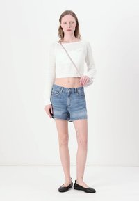 Hvit, strikket, langermet crop-top kombinert med lyseblå denimshorts. Modell iført svarte flate sko, står mot en nøytral bakgrunn.