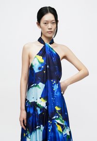 Sininen halterneck-mekko, jossa on abstrakteja monivärisiä kuvioita vihreänä, keltaisena ja valkoisena. Valmistettu sileästä, kevyestä kankaasta, jossa on kerättyjä yksityiskohtia.