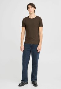 T-shirt marron à manches courtes en tissu doux, associé à un jean bleu foncé. Le modèle se tient debout en chaussures noires sur un fond uni.