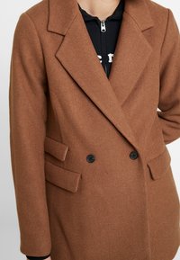 Abrigo marrón de doble botonadura con un acabado texturizado. Presenta amplios solapas, dos botones negros y bolsillos laterales con detalles de solapa.