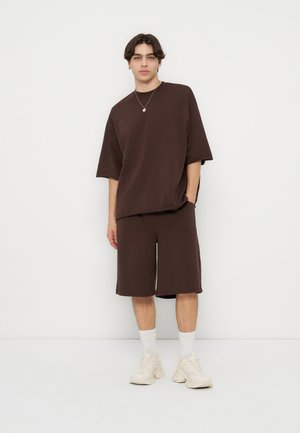 Joven de pie con camiseta de gran tamaño marrón oscuro y pantalones cortos largos a juego, calcetines blancos y zapatillas blancas gruesas sobre un fondo liso.