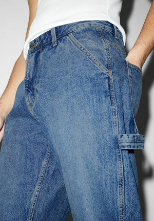 Personne portant un jean en denim bleu et un haut blanc sans manches, main dans la poche, avec des passants de ceinture visibles et un détail de boucle latérale.