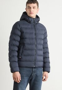 Mornarica puffer jakna s kapuljačom, vertikalnim kvadratnim uzorkom, prednjim zatvaračem i suptilnim logotipom s lijeve strane.