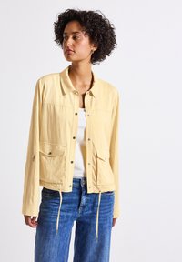 Giacca leggera gialla con collo a camicia, chiusura a bottoni a pressione, due tasche frontali e orlo con cordino, abbinata a jeans blu.
