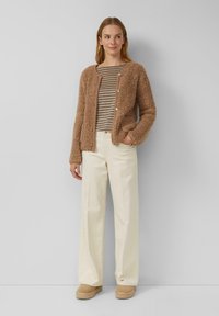 Frau trägt eine braune, flauschige, knöpfbare Jacke, ein gestreiftes Shirt, cremefarbene weit geschnittene Hose und beige Plateauschuhe, steht mit einer Hand in der Tasche.