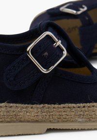 Navy canvas shoe with espadrille-style woven jute sole, featuring a silver buckle and textured strap design. Flat sole, casual cut.