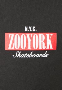 Sort stof har et iøjnefaldende rød- og hvidt grafisk design med teksten "N.Y.C. ZOOYORK Skateboards", sat mod en struktureret baggrund.