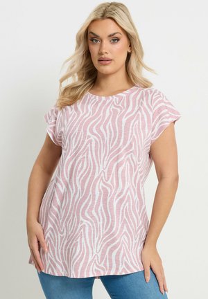 Blondynka w różowo-białym topie w paski zebry z krótkim rękawem i niebieskich dżinsach, pozująca z rękami blisko bioder.