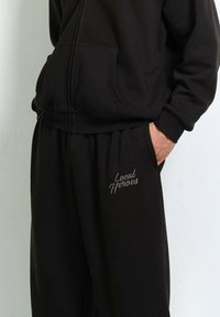 Person in einem schwarzen Hoodie und passender Jogginghose mit dem Aufdruck "Local Heroes" auf dem linken Oberschenkel, mit einer Hand in der Tasche.