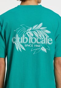 Tamsiai mėlynas medvilninis marškinėliai su balta lapų raštu ir tekstu "club locale" su "SINCE 1960" žemiau. Trumpomis rankovėmis, laisvo kirpimo.