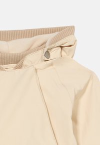 Μπεζ μπουφάν με ριμπ ύφασμα στη φόδρα του γιακά και κλείσιμο με κουμπιά κοντά στον λαιμό, εμφανιζόμενο από την περιοχή του πάνω ώμου.