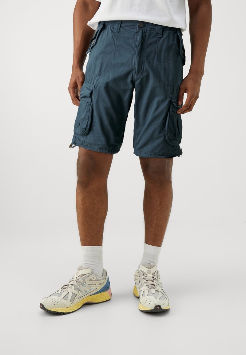 Homme portant un short cargo bleu foncé avec des poches latérales, des chaussettes blanches et des baskets de sport multicolores sur un fond clair uni.