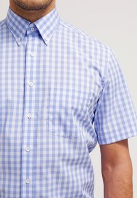 Vaaleansininen gingham-kuvioitu lyhythihainen paita, jossa on napillinen kaulus, etutasku ja valkoiset napit rapeasta puuvillakankaasta.