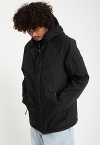 Homme aux cheveux bouclés portant une veste noire à capuche avec des poches zippées, regardant vers le bas, mains dans les poches, avec un jean bleu clair.