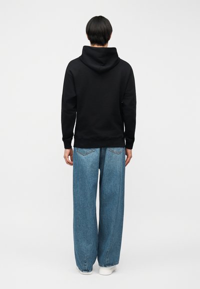 Personne debout de dos portant un sweat à capuche noir uni, un jean bleu ample et des baskets blanches sur un fond blanc.