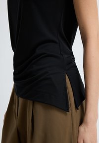 Černé, lehké tričko s bočními rozparky a hladkou texturou. V kombinaci s hnědými, vysokými kalhotami, které zdůrazňují uvolněný střih a splývání.
