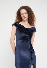 Σκούρο μπλε φόρεμα χωρίς τους ώμους με ντεκολτέ με draped ύφασμα, λεπτομέρεια με βολάν και πλαϊνή σχισμή. Κατασκευασμένο από λείο, γυαλιστερό ύφασμα που αντανακλά το φως.
