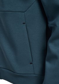 Felpa blu scuro con superficie testurizzata, caratterizzata da una tasca laterale con un accento nero. Linee pulite e design minimalista.