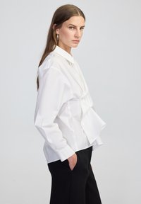 White button-up shirt with a foldover design, long sleeves, and a structured collar. Worn with black tailored pants, showcasing clean lines.