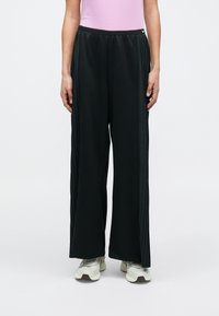 Czarne sportowe spodnie z bocznymi plisami, luźnym krojem, elastycznym pasem i detalem z logo, zestawione z jasnymi butami sportowymi.