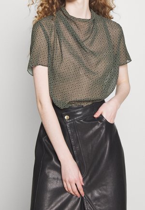 Vrouw die een doorschijnend patroonachtig kort mouwtopje draagt, ingestopt in een hoge taille zwarte leren rok, met hand in de zak tegen een effen achtergrond.