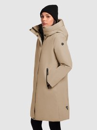 Beige lang frakke med oversized hætte, sort hardware og sidelommer. Båret over en sort top og hue med et logo på kanten.