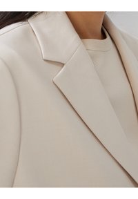 Světle béžový blazer s klopovým límcem, strukturovanou látkou a přiléhavým střihem, vrstvený přes krémový pletený top.