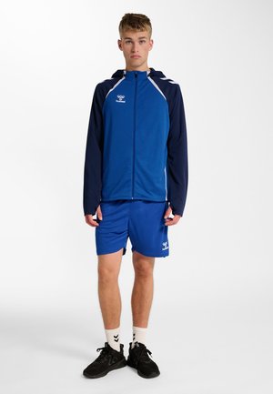 Jonge man staat en draagt een blauwe en marineblauwe sportjas met bijpassende blauwe korte broek, witte sokken en zwarte sportschoenen op een witte achtergrond.