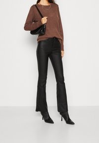 Pull en maille marron, pantalon en cuir noir évasé, bottines à talons noires et petit sac à main noir avec bandoulière en chaîne.
